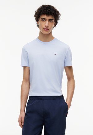 Giovane uomo con i capelli ricci che indossa una t-shirt azzurro chiaro e pantaloni blu scuro, in piedi con una mano in tasca su sfondo bianco.