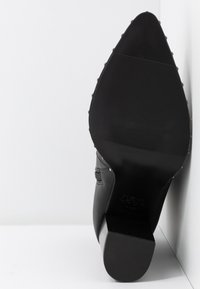 Scarpa nera con tacco alto e punta affusolata, suola in gomma testurizzata e tomaia in pelle liscia. Presenta borchie decorative in metallo lungo il retro.