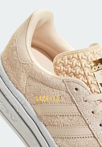 Beige suede sneaker featuring three parallel stripes, embossed accent on the tongue, gold "GAZELLE" text, and a textured sole.
