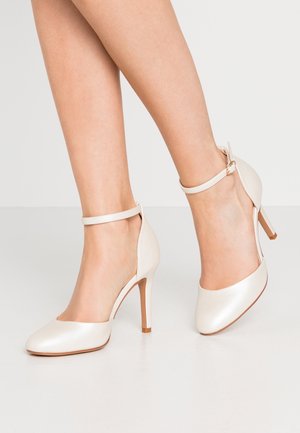 Jambes de femme portant des escarpins ivoire fermés à bout rond avec des brides à la cheville, posées sur une surface blanche lisse sur un fond uni.