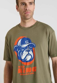 Hombre con una camiseta verde oliva que tiene un gráfico de un bulldog con una gorra azul dentro de un óvalo naranja y el texto "EL REY nacido para ganar" debajo.