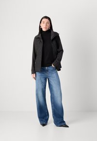 Een jonge vrouw staat zelfverzekerd in een zwarte vest over een zwart top en brede, blauwe jeans, met oorbellen en puntige schoenen.