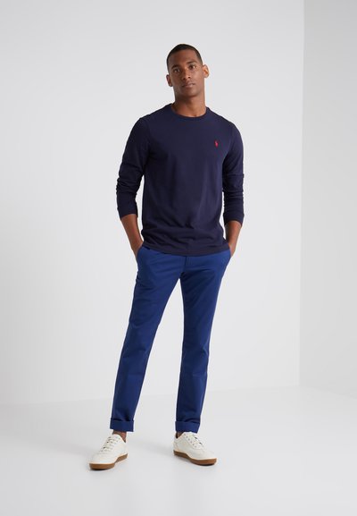 Marineblauwe longsleeve shirt met een geribbelde halslijn, blauwe chinos en witte sneakers met beige accenten, staand tegen een lichte achtergrond.