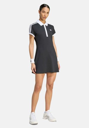 Frau trägt ein schwarz-weißes Adidas Tenniskleid, weiße Handgelenksbänder und weiße Turnschuhe, steht mit entspannten Armen vor einem einfarbigen Hintergrund.