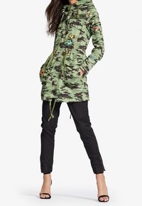 Kamouflageparka i grön tyg med dragsko i huven, två framfickor och blommig broderi. Matchas med svarta byxor och klackar.