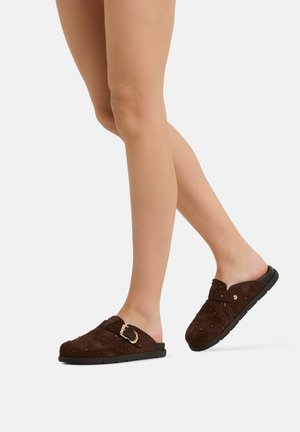 Beine, die dunkelbraune Slip-On-Mules mit Schnallen- und Nieten-Details tragen, stehen vor einem einfarbigen hellen Hintergrund.