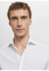 Biała koszula zapinana na guziki z klasycznym kołnierzykiem, o gładkiej fakturze i minimalistycznym designie, noszona przez modelkę na jasnym tle.