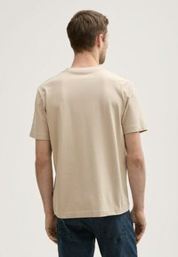 Beige, rövid ujjú póló sima anyagból, kerek nyakkivágással és egyenes szabással. A hátsó része minimális részletezéssel és nyomatok nélkül készült.