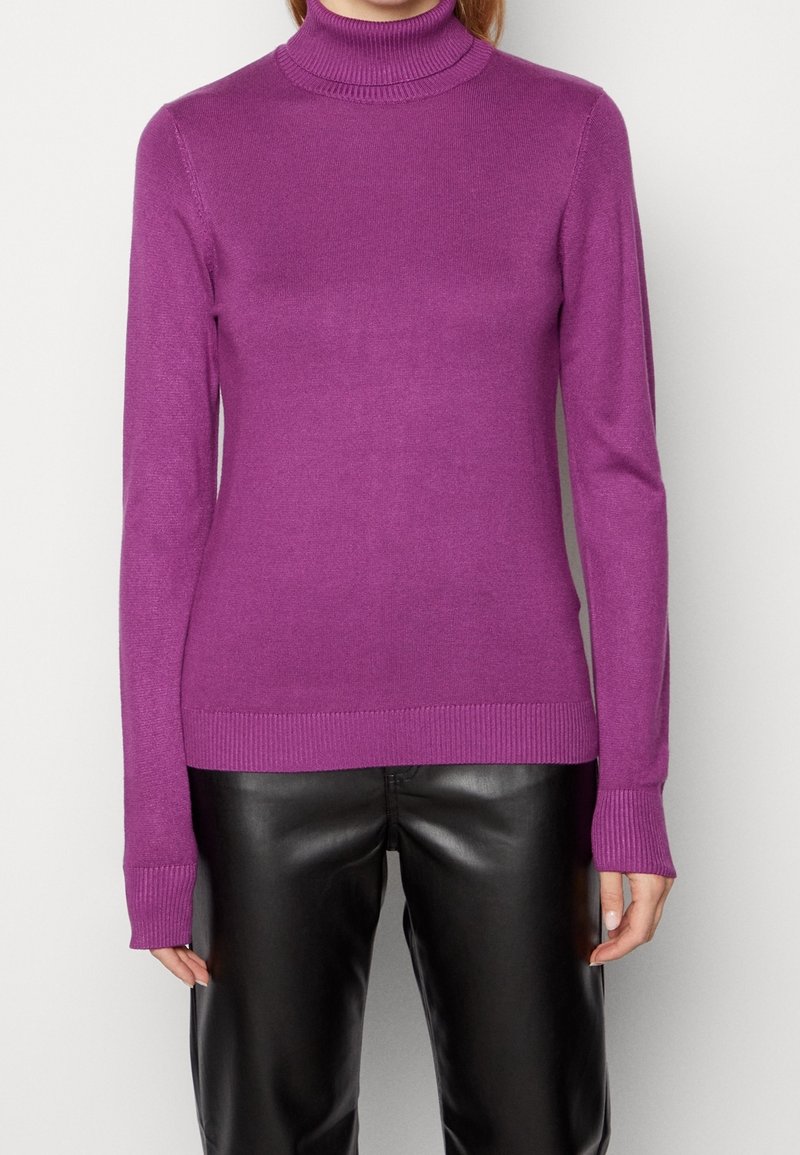 Femme portant un pull à col roulé violet à manches longues et un pantalon en cuir noir, debout devant un fond uni clair.