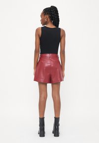 Top nero senza maniche abbinato a pantaloni corti in pelle bordeaux lucida a vita alta. Il modello indossa stivaletti neri alla caviglia. Texture liscia, design aderente.