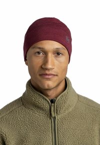 Young man wearing a maroon beanie and a beige fleece jacket with a high collar, facing forward against a white background.