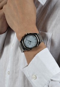 Silberner Metallarmbanduhr mit achteckigem Gehäuse, strukturiertem hellblauen Zifferblatt, silbernen Zeigern und metallischem Armband, getragen an einem Handgelenk mit einem weißen Hemd.