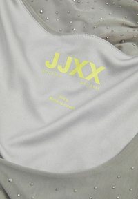 Sivi bez rukava s glatkom teksturom, s detaljima od srebrnih zakovica i hrabrim žutim "JJXX" brendom. Veličina S, proizvedeno u Kini.