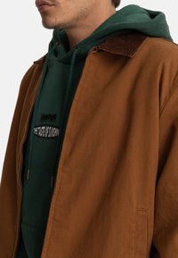 Ruskea puuvillatakke corduroykankaalla vuoratulla kauluksella, päällä vihreä huppari, jossa on logo ja kiristysnauhat. Sileä pinta, rento istuvuus.