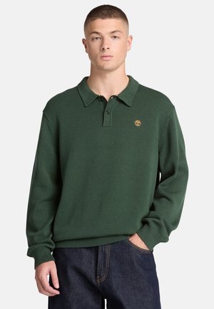 Jonge man draagt een donkergroen gebreide polo met lange mouwen en een klein geel logo op de linkerborst, gecombineerd met donkerblauwe spijkerbroek.