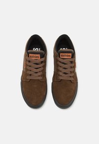 Sapatilhas de camurça castanha com forro preto, atacadores castanhos e logótipos laranja da Etnies na lingueta. Sola de borracha com uma textura subtil.