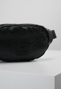 Czarna nylonowa nerka o pikowanej teksturze, z przednią kieszenią na zamek błyskawiczny i plecionym paskiem. Zawiera kwadratową naszywkę z logo z boku.