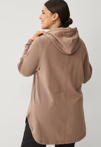 Brown hooded cardigan with long sleeves, soft texture, a curved hem, and side slits. No visible patterns or hardware accents. Black pants.