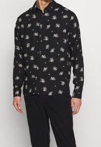 Camicia button-up nera con motivi floreali bianchi, maniche lunghe e colletto a punta, realizzata in tessuto leggero.