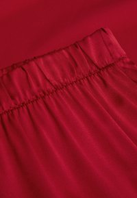 Rode stof met een gladde textuur, een geplooide elastische tailleband en een subtiele glans, wat zorgt voor een vloeiende uitstraling.