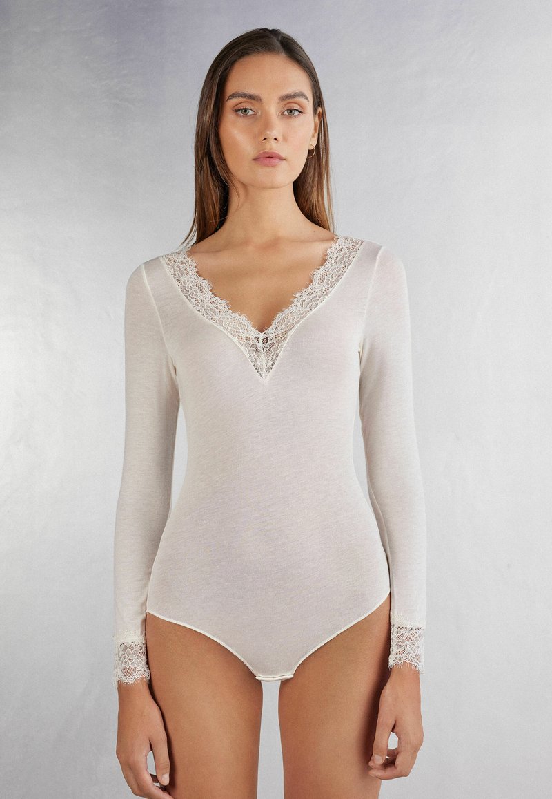 Bodysuit de manga larga en suave tela color blanco roto, que presenta un escote en V profundo con acabado de encaje y detalles de encaje en los bordes de los puños. Textura suave.