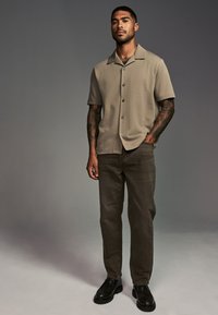 Beige overhemd met korte mouwen en een spread collar, gecombineerd met donkergroene broek en zwarte chunky schoenen, voor een neutrale achtergrond.