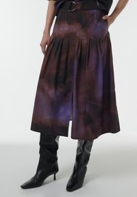 Una falda plisada morada y marrón con una abertura lateral, fabricada en un tejido ligero, combinada con botas de cuero negras de tacón alto y puntera afilada hasta la rodilla.