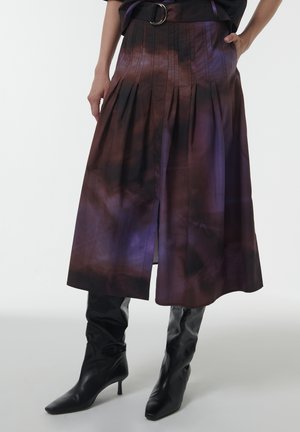 Une jupe plissée violet et marron avec une fente sur le côté, fabriquée en tissu léger, associée à des bottes noires en cuir à talons aiguilles jusqu'aux genoux.