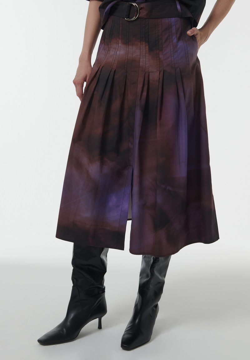 Una falda plisada morada y marrón con una abertura lateral, fabricada en un tejido ligero, combinada con botas de cuero negras de tacón alto y puntera afilada hasta la rodilla.