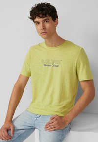 Żółta bawełniana koszulka z okrągłym dekoltem i krótkimi rękawami. Z przodu znajduje się logo "S.OLIVER Elevated Casual" w ciemnoniebieskim kolorze.