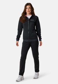 Black athletic zip-up jacket with a smooth texture, paired with matching pants featuring a logo detail on the leg. White sneakers complete the look.