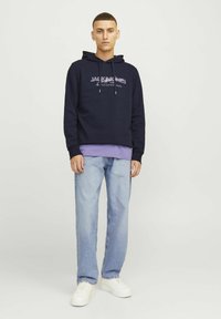 Sudadera de algodón marina con cordón, camiseta de cuello redondo morada debajo, jeans de mezclilla azul claro y zapatillas blancas, de pie sobre un fondo liso.