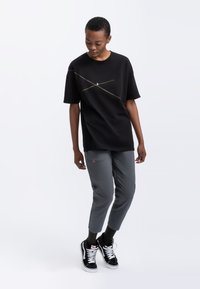 T-shirt en coton noir à manches courtes avec un motif de ligne diagonale dorée. Associé à un pantalon gris ajusté et des baskets noires.