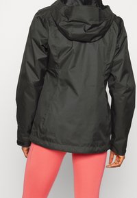 Veste imperméable noire avec capuche, présentant une texture lisse, des poches avant et des poignets ajustables, portée sur des leggings roses.