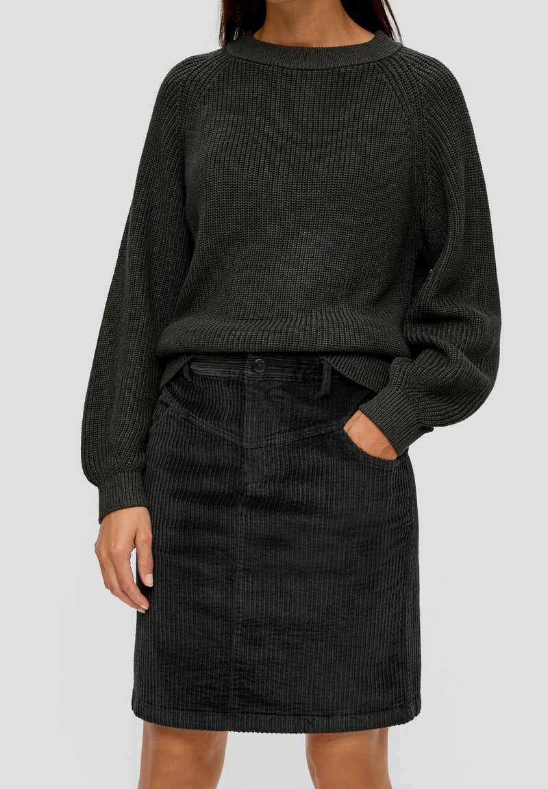Femme portant un pull en maille foncé avec des manches ballon rentrée dans une jupe en velours côtelé noire, debout avec une main dans la poche.