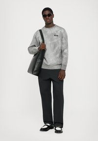 Hellgrauer Batik-Sweatshirt mit Logo, schwarze weite Hose, schwarze Tote Bag und schwarze Sonnenbrille. Die Schuhe haben ein zweifarbige Design.