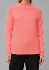 Korallfarbener Strickpullover mit strukturiertem Finish, hochgeschlossenem Ausschnitt und langen Ärmeln, kombiniert mit schwarzen Jogginghosen mit Kordelzug.