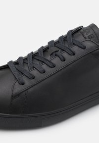 Black leather sneaker with flat black laces, smooth texture, low-profile design, and rubber outsole with grip pattern.
