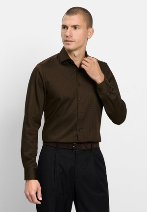 Man die zijn kraag aanpast, draagt een donkerbruine lange mouwen shirt en zwarte broek met een bijpassende riem tegen een effen achtergrond.