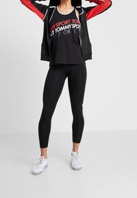 Donna che indossa leggings neri Tommy Sport, canotta nera con logo rosso, giacca nera con maniche rosse e sneakers bianche, in piedi con le braccia alzate.