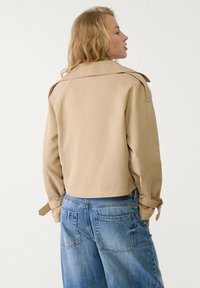Beige Z cropped Jacke mit weitem Kragen und Bundbändern, kombiniert mit locker sitzenden blauen Jeans aus Denim mit großen Gesäßtaschen.