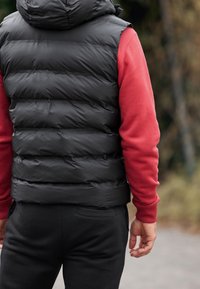 Schwarze, wattierte Weste mit Kapuze, kombiniert mit einem roten Langarmshirt. Die Weste zeichnet sich durch horizontale Steppnähte und eine körpernahe Passform aus.
