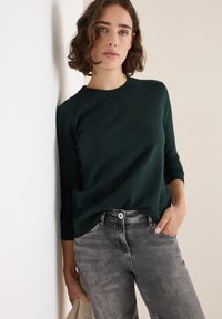 Dunkelgrüner Pullover mit Rundhalsausschnitt und langen Ärmeln, kombiniert mit hellgrauen Jeans mit Knopfverschluss und Fronttaschen.