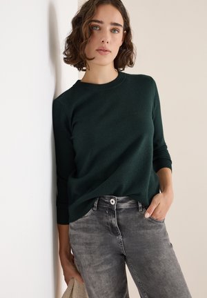 Suéter verde oscuro con cuello redondo y mangas largas, combinado con jeans gris claro que presentan un cierre de botón y bolsillos frontales.