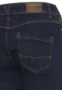 Blugi din denim albastru închis, văzuți din spate, cu cusături portocalii și o etichetă din piele pe talie pe care este inscripționat "Pulz Jeans".