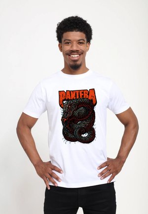Lächelnder Mann mit lockigem Haar, der ein weißes T-Shirt mit einer gewundenen schwarzen Schlange und dem Wort "PANTERA" in fetten roten Buchstaben trägt.