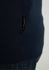 Ourlet de pull en maille bleu foncé avec une étiquette en tissu noire indiquant "CHASIN' 7 92 1059" porté sur un jean en denim foncé.