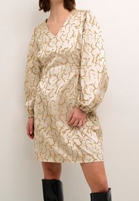 Beige klänning med v-ringning, långa puffärmar och ett gyllene abstrakt mönster. Har samlade manschetter och en avslappnad passform. Bärs med svarta knähöga stövlar.