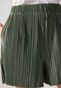 Shorts plissés en tissu vert foncé avec une surface texturée. Dotés d'une taille élastique et de poches latérales pour plus de fonctionnalité.