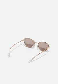 Óculos de sol estilo gato com lentes castanhas, armação fina em metal rosa dourado e pontas em plástico transparente nas hastes. Design minimalista.